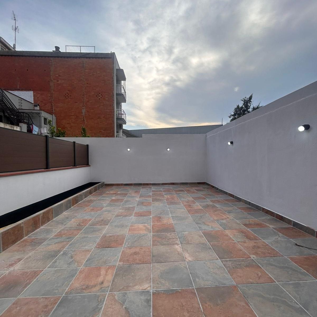 La misma terraza reformada: gres en tonos tierra, frente de lamas y apliques